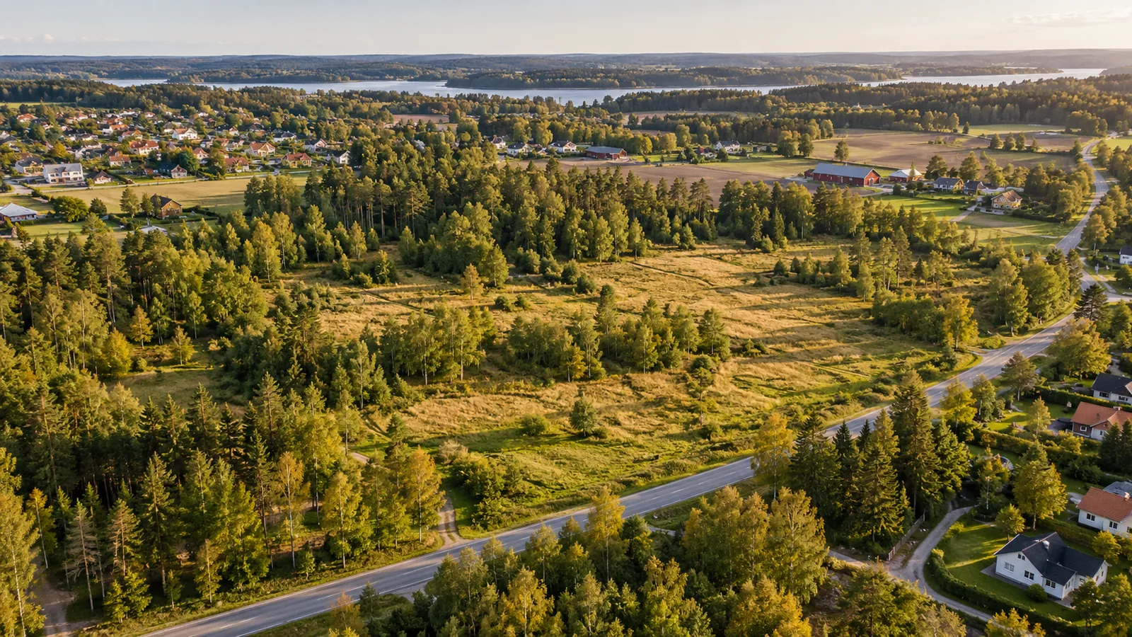 Obebyggt markområde med äng och skogsdungar intill en väg och ett villaområde